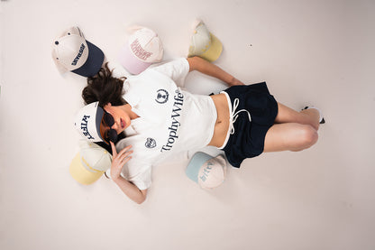 Person wearing a white polo with 'Trophy Wife' text, surrounded by multiple hats on a light gray background including husband and wifey hats