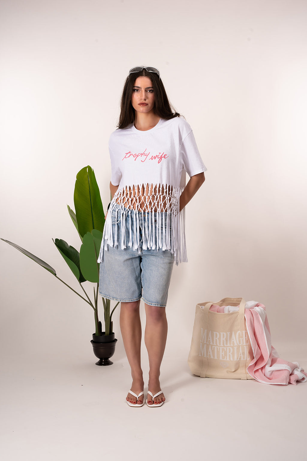 Woman wearing a white t-shirt with text and a fringed top, standing next to a plant and a bag with text on a plain background