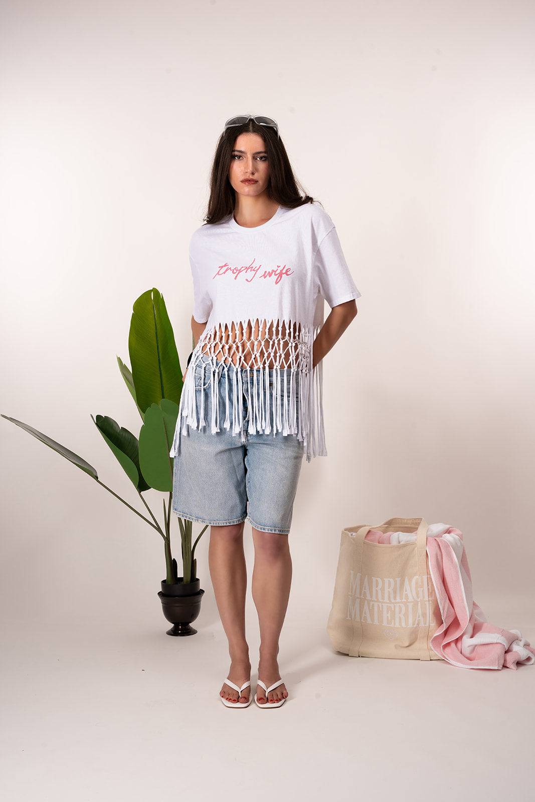 Woman wearing a white t-shirt with text and a fringed top, standing next to a plant and a bag with text on a plain background
